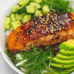 Salmon Teriyaki topped on rice with slice avocado and cucumber salad on the side in a bowl.