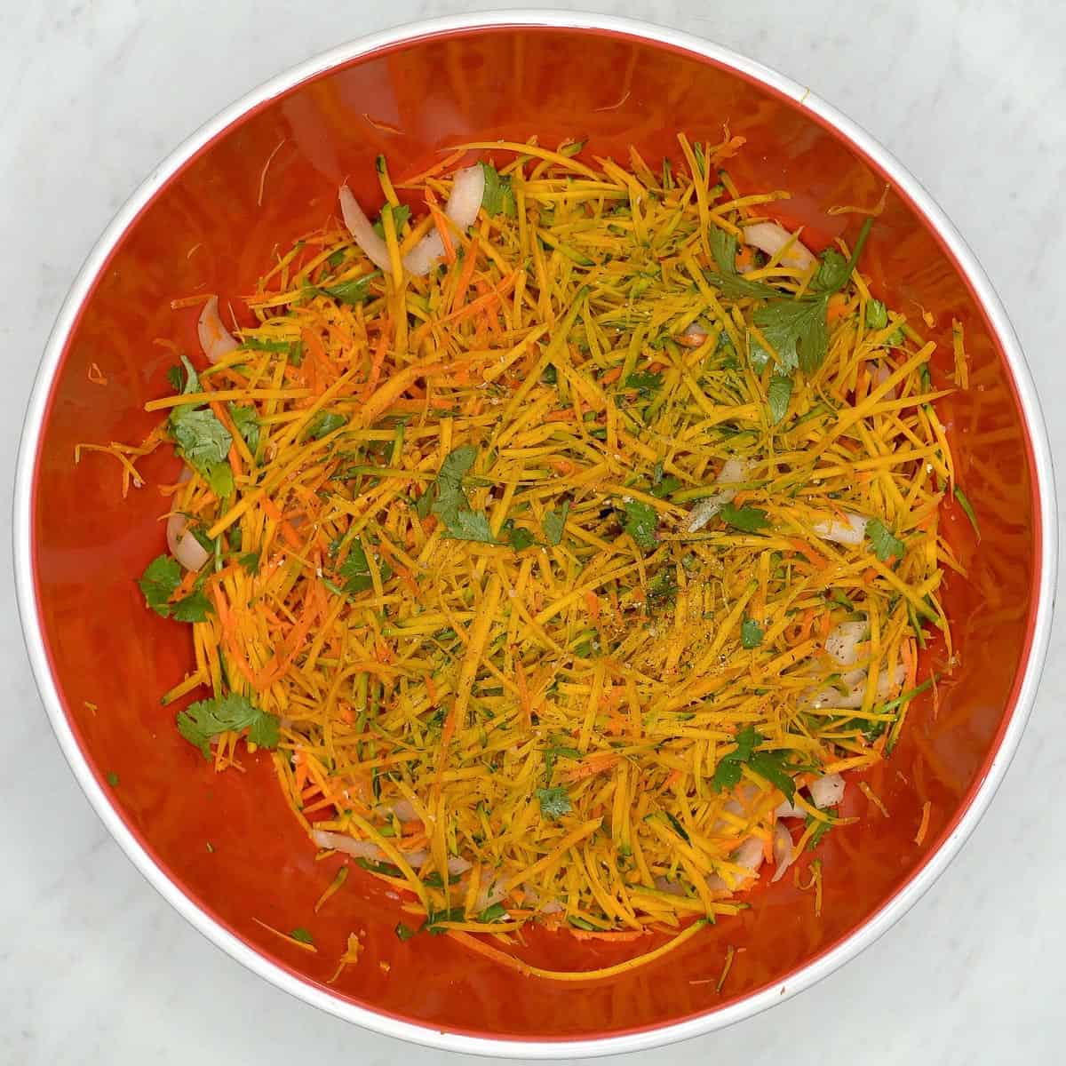 A red bowl filled with julienned squash and carrots, onion, cilantro, salt and pepper.