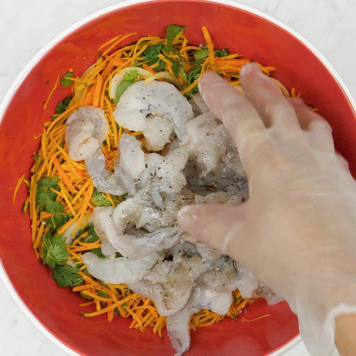 A red bowl with seasoned shrimp and vegetables about to be mixed by hand.