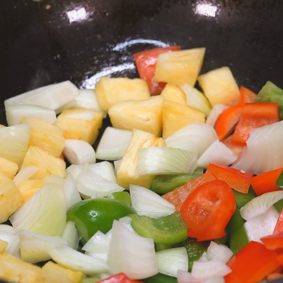 Pineapple, onions, green bell peppers, and red bell peppers spread out in a wok.
