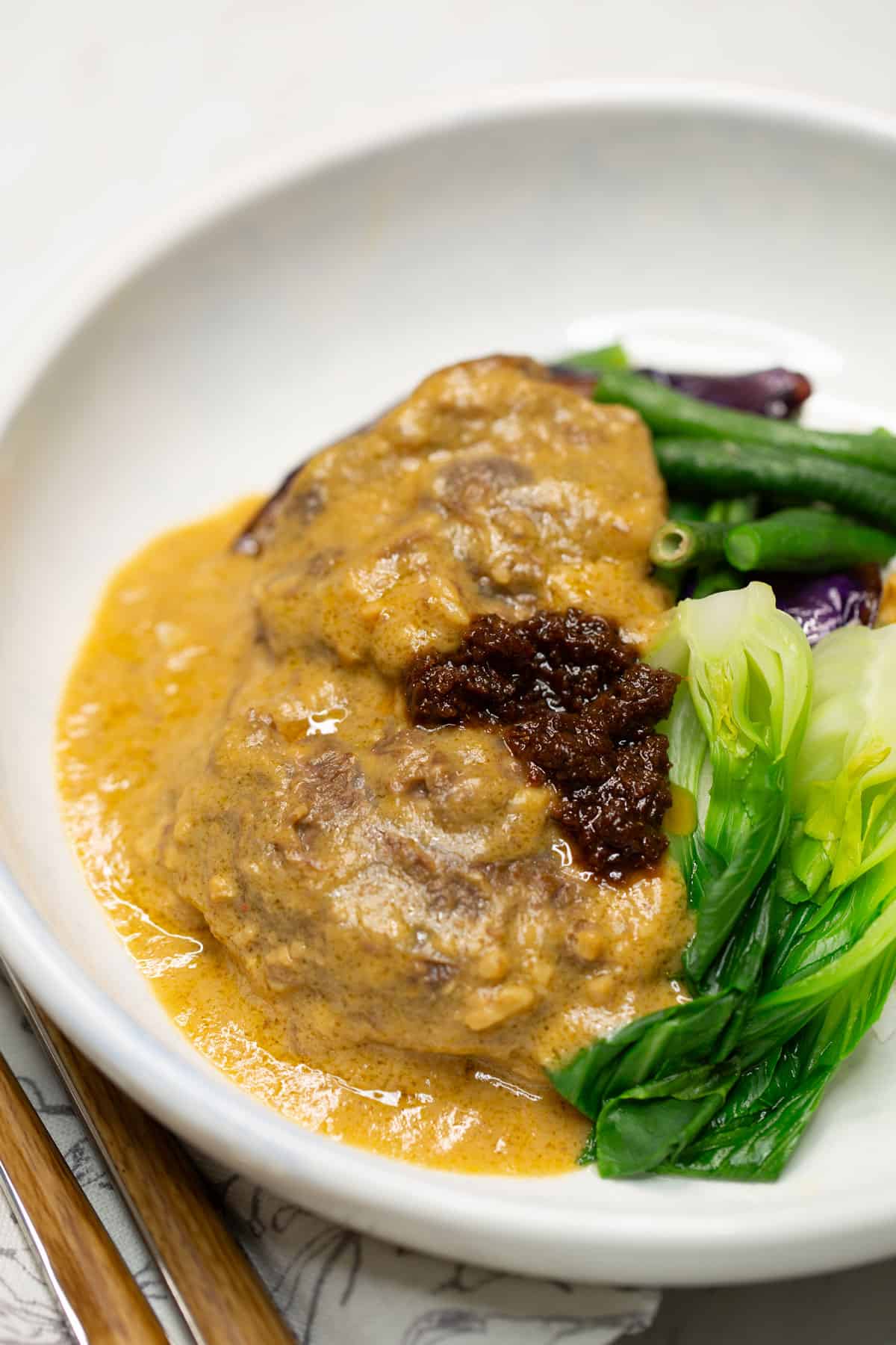 Kare Kare with beef shank served with rice on a plate with a side of sauteed shrimp paste, more rice, a fork, knife, and napkin.