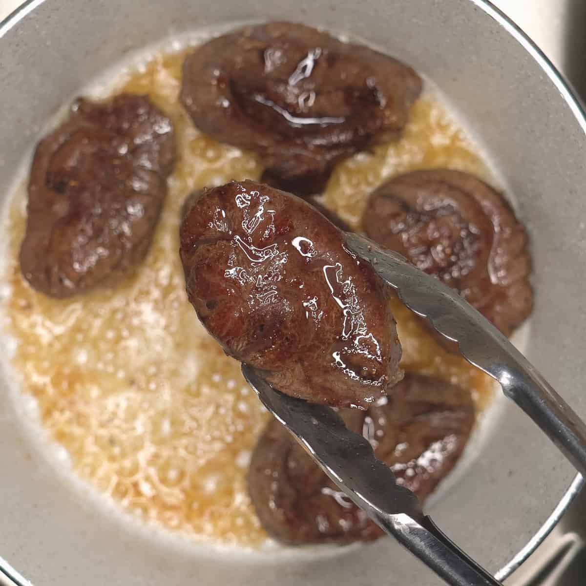 Seared pieces of meat in oil and a piece being lifted off the pot with tongs.
