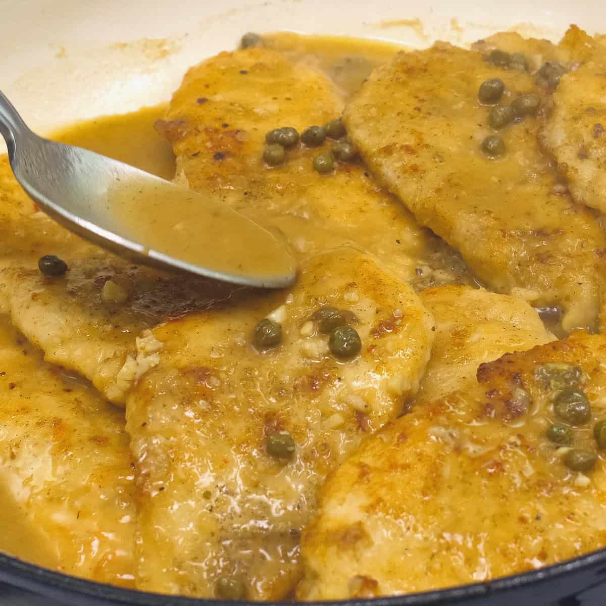 Scooping the sauce over the chicken in the pan with a spoon.