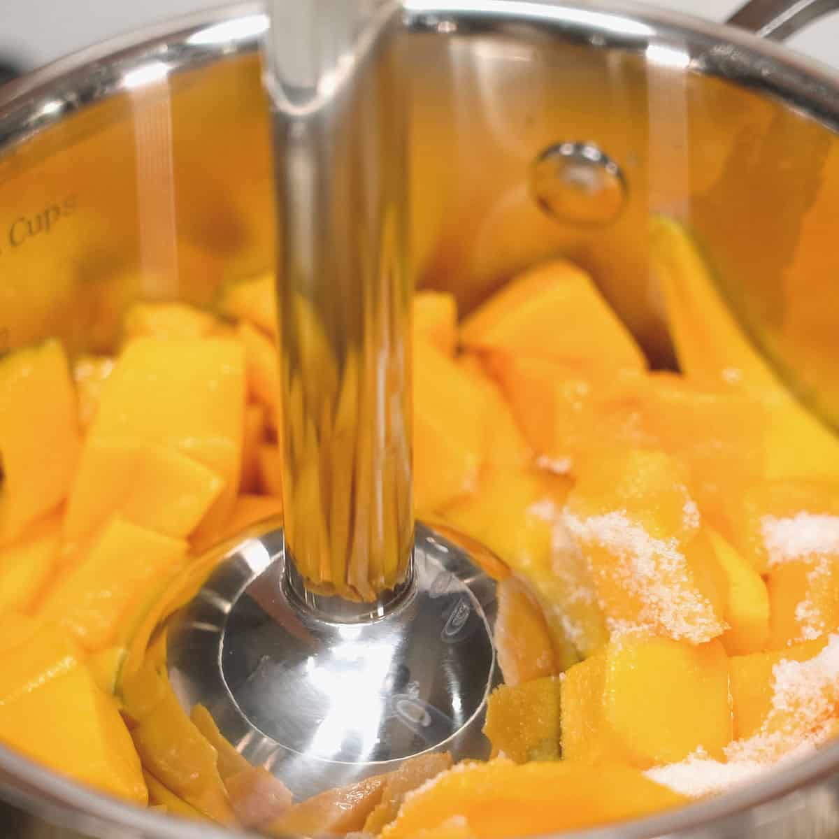 Blending mangoes and sugar using an immersion blender in a saucepan.