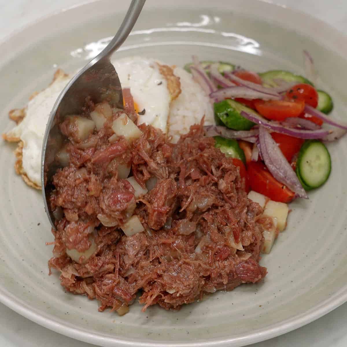 Adding ginisang corned beef to a plate with rice, fried egg, and salad.