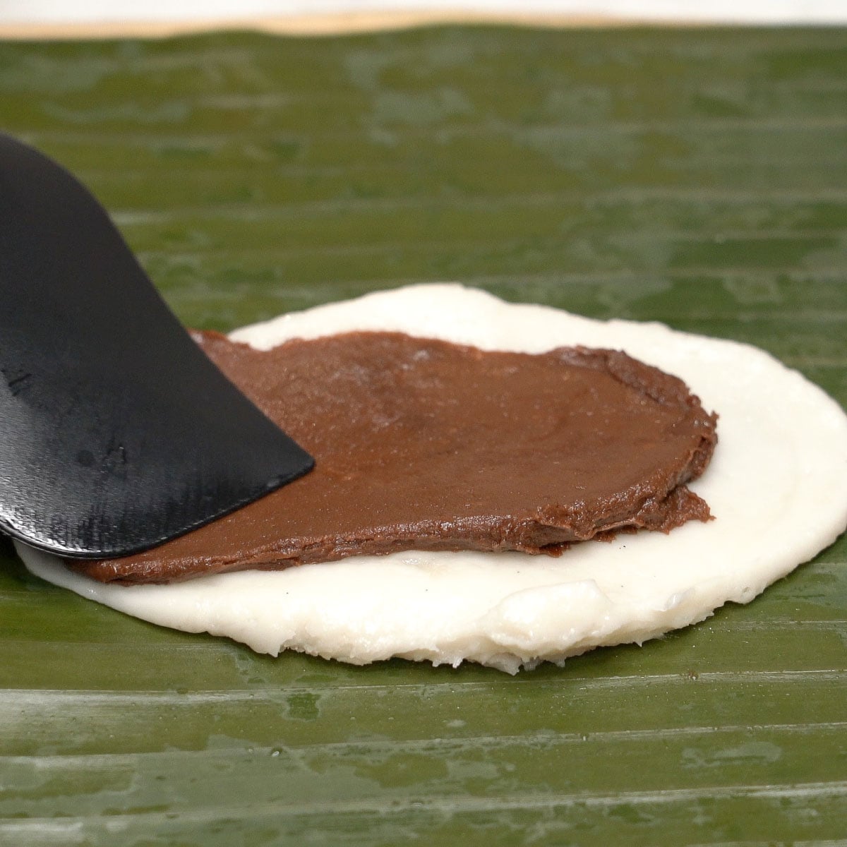 Spreading chocolate mixture on top of white mixture on a banana leaf.
