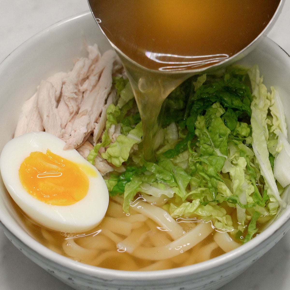 Pouring the broth into the bowl of noodles, chicken, boiled egg, and napa cabbage.