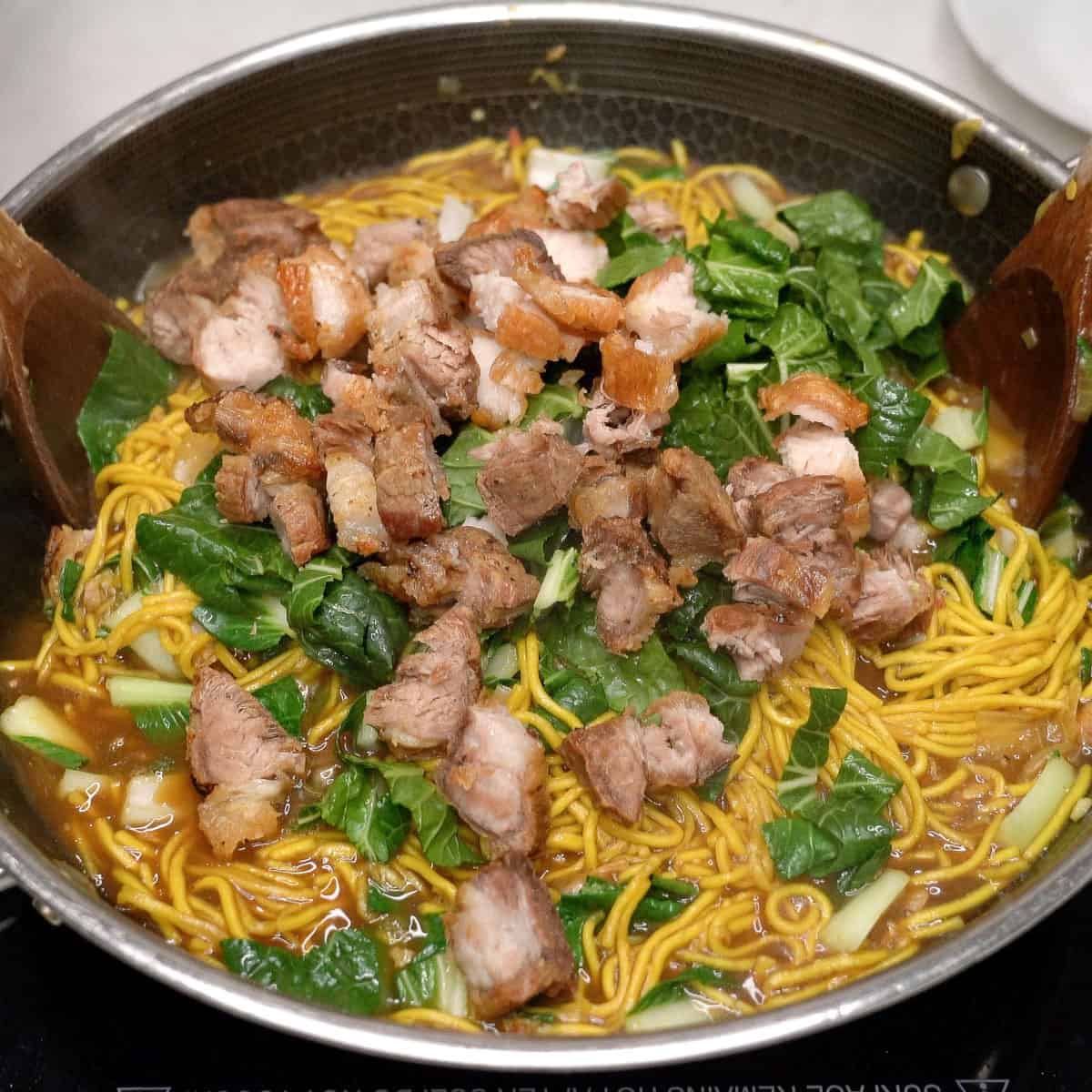 Tossing pancit bato noodles with pechay and crispy pork in a wok.