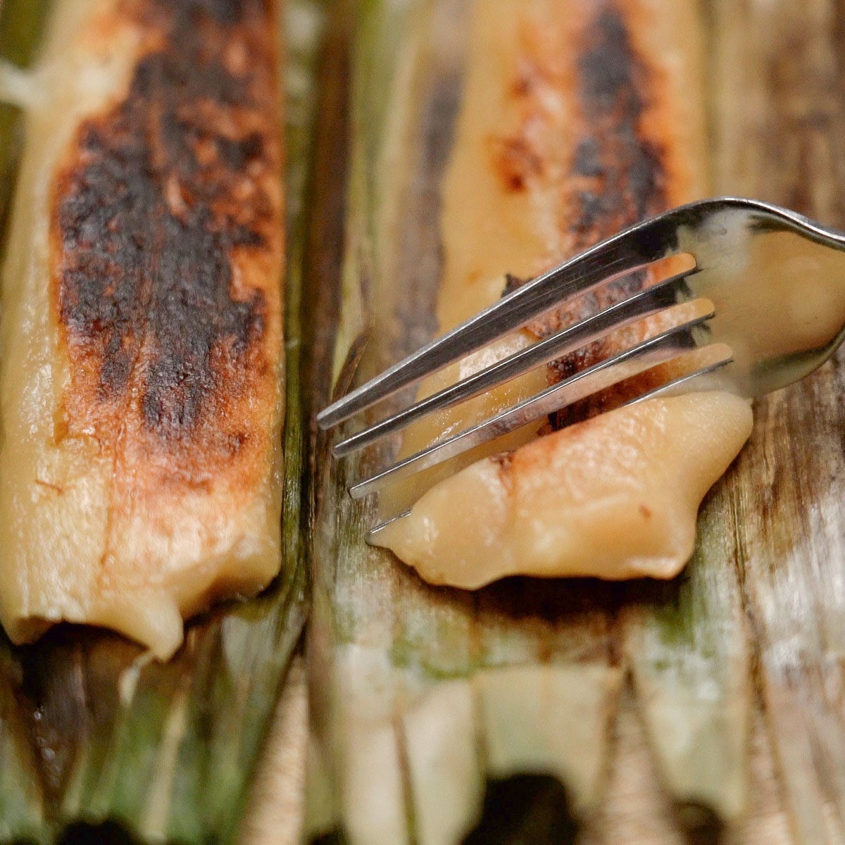 Fork cutting into tupig with charred top on banana leaves.