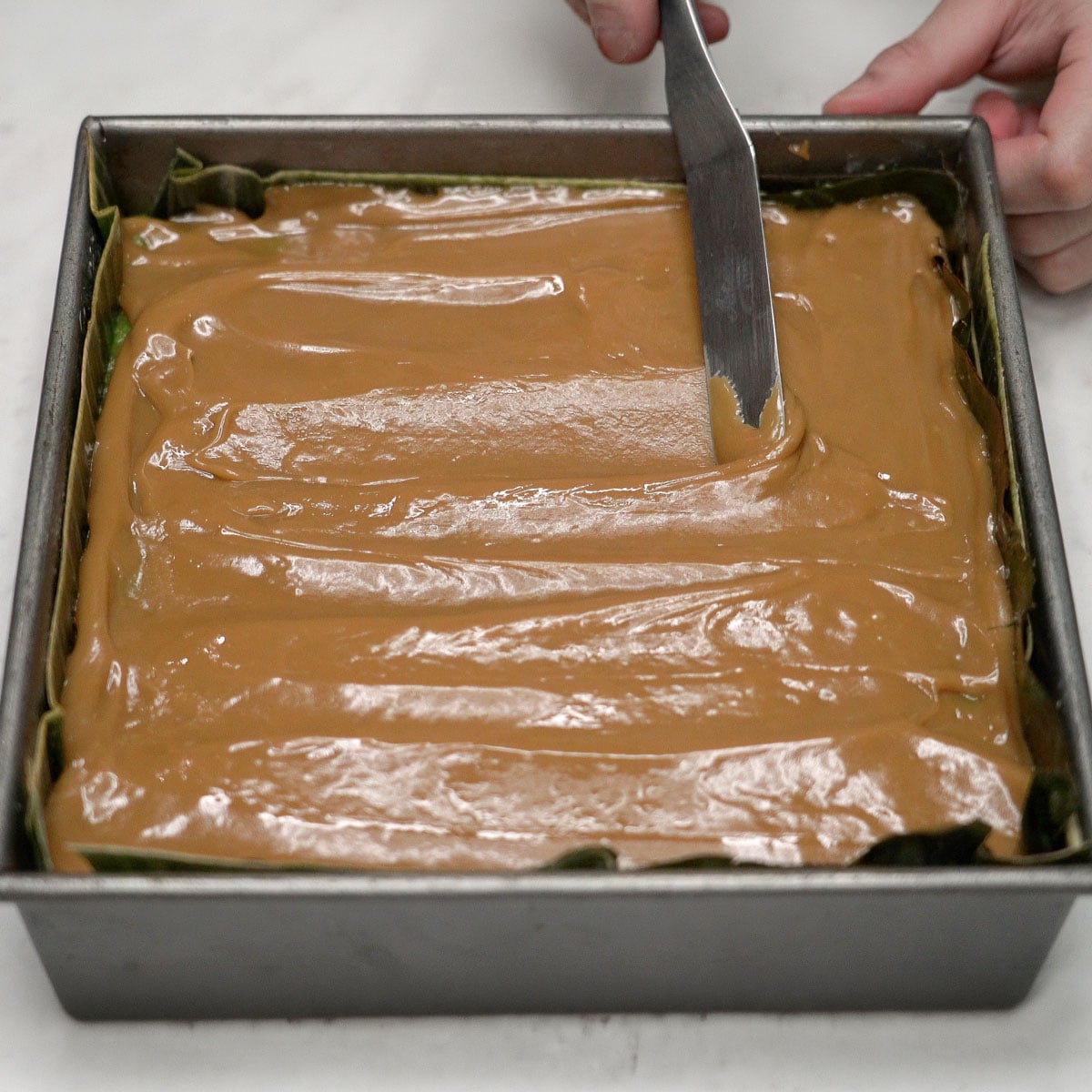 Spreading latik sauce on the bibingkang pinipig mixture in the pan.