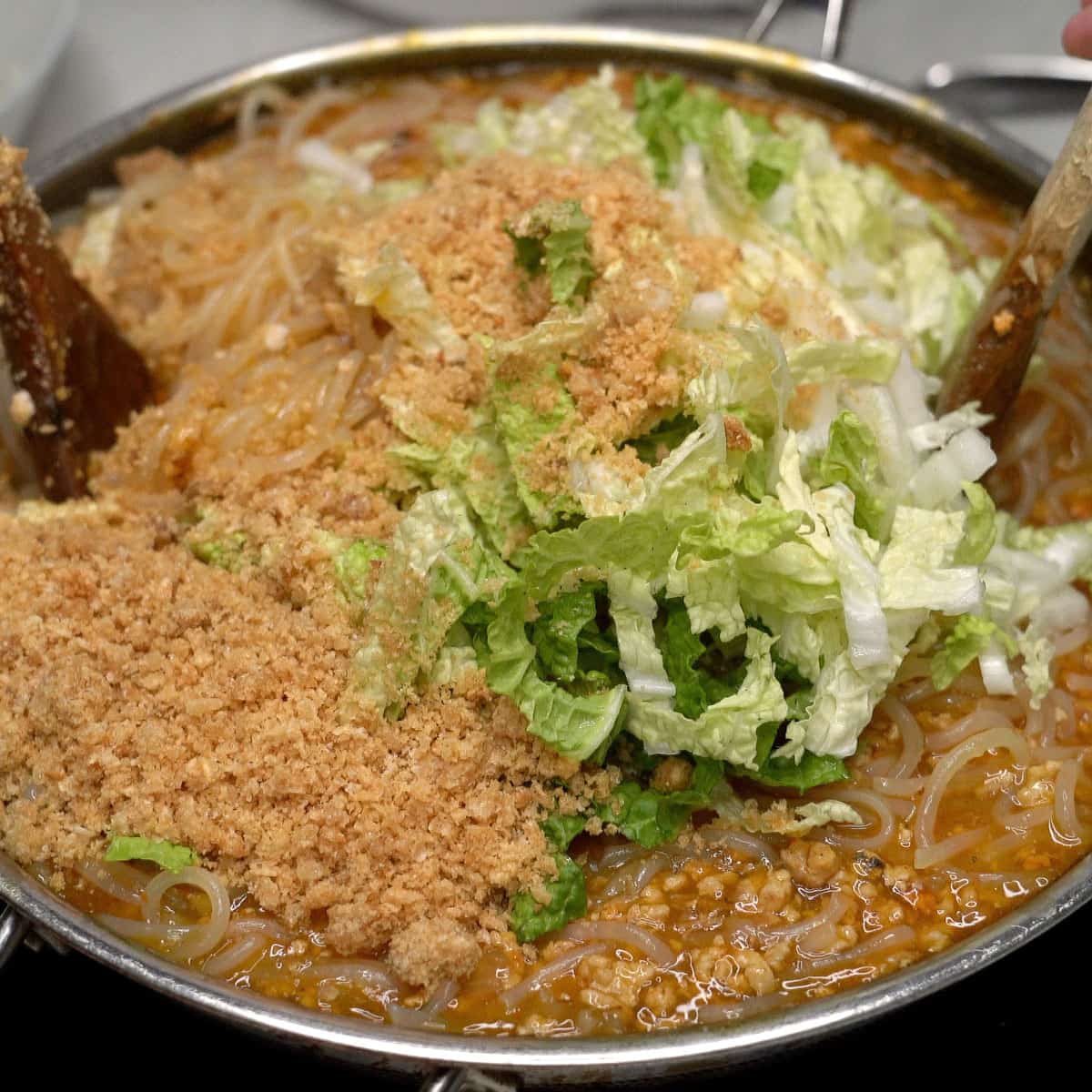 Crushed chicharon and napa cabbage added to the pancit malabon in the wok.