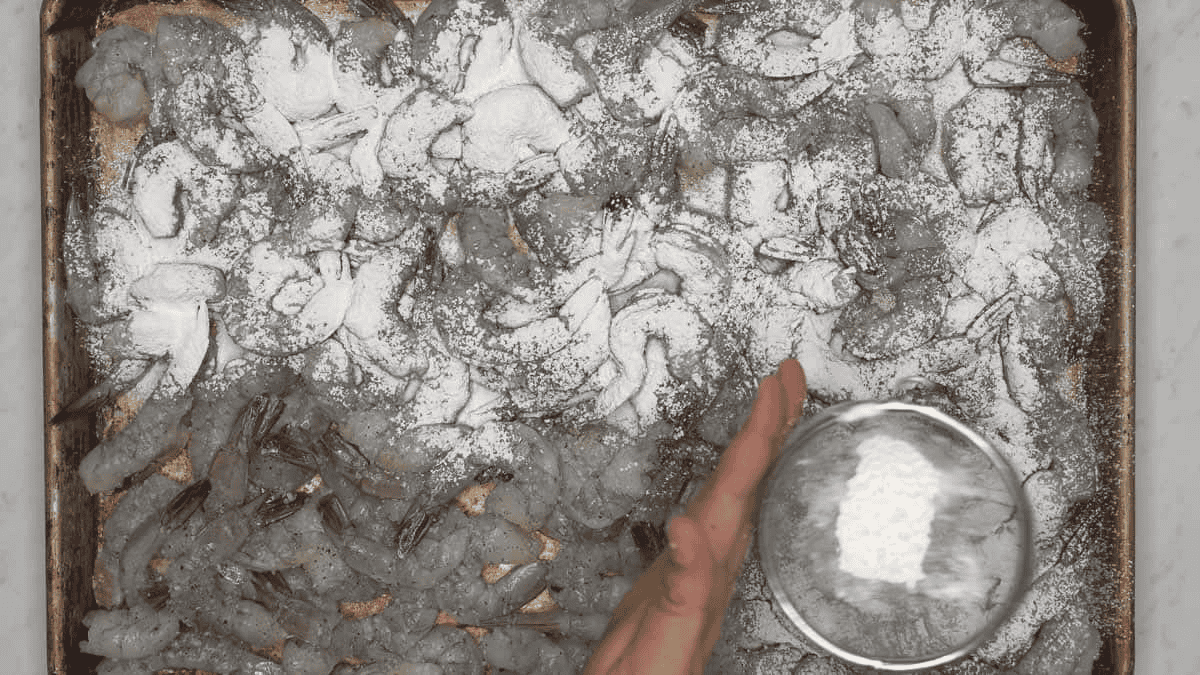 Adding cornstarch to shrimps spread out on a baking sheet.