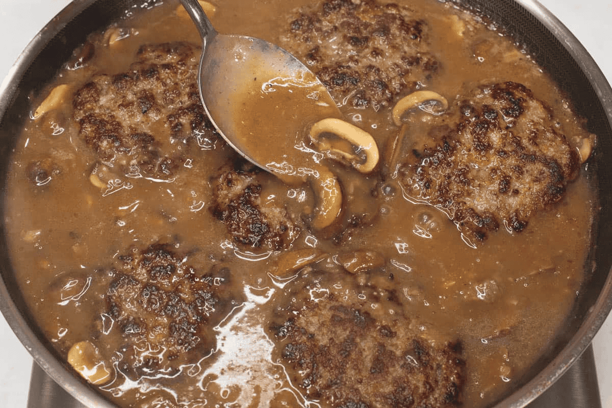 Spooning the mushroom gravy over the burger patties in the skillet.