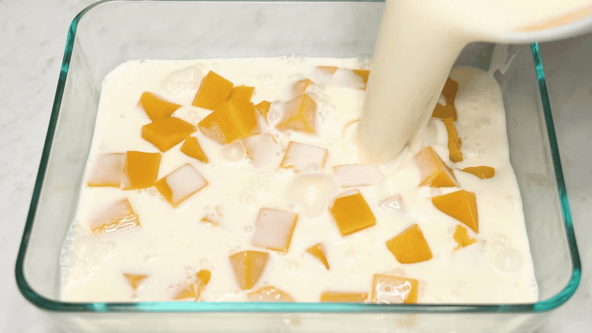 Pouring the cream gelatin mixture over the mango cubes in the container.