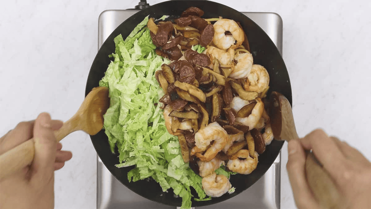 Mixing the cabbage, mushrooms, sausage, and shrimp with the noodles in the wok.