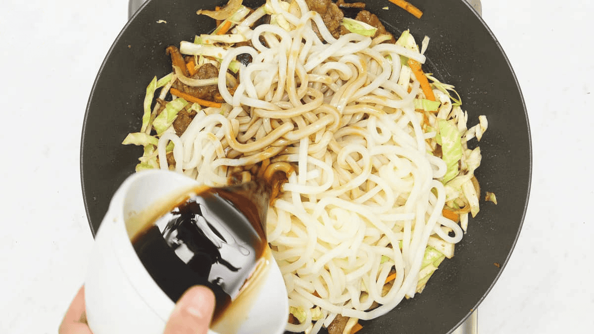 Adding the sauce to the udon noodles in the wok.