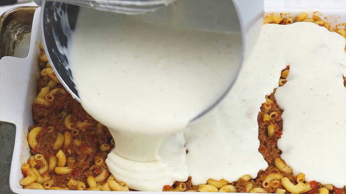 Adding the bechamel sauce on top of the macaroni with meat sauce in the baking dish.