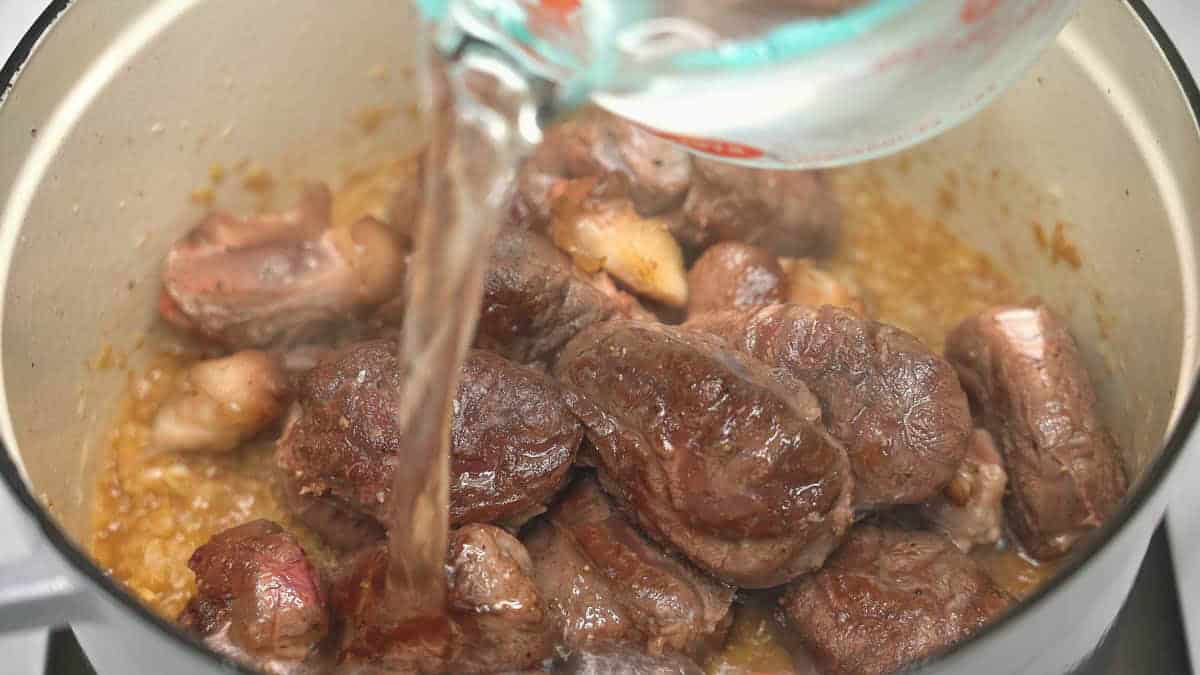 Adding water to the seared meat and aromatics in a Dutch oven.