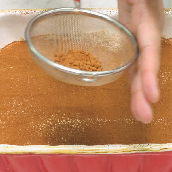 Dusting cocoa powder on the layer of mascarpone mixture using a small sieve. Spreading a layer of mascarpone mixture with a spoon on top of the ladyfingers arranged in the baking dish.