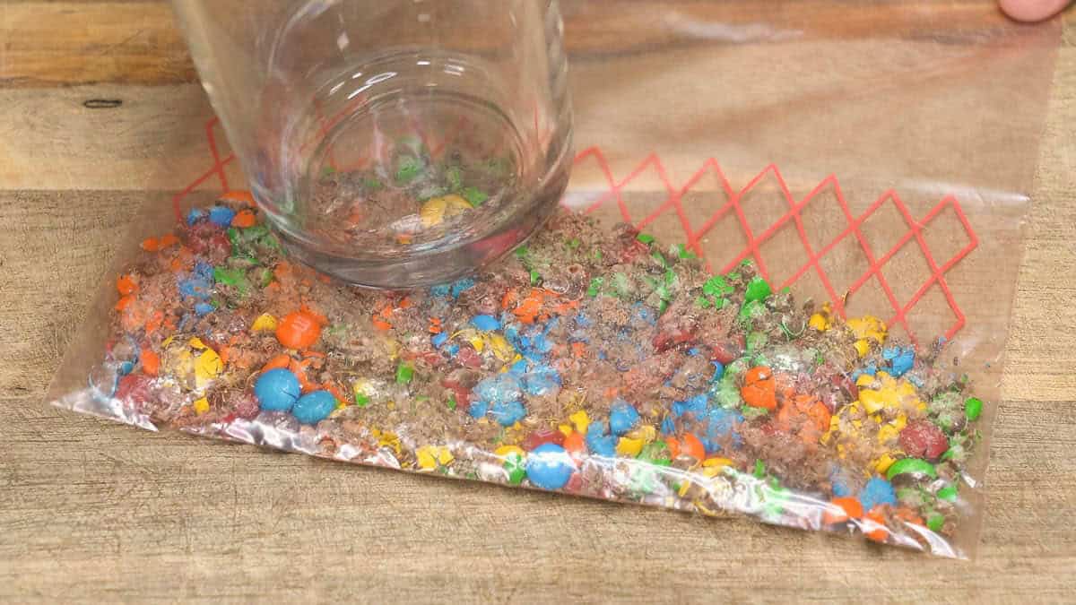 Using the bottom of a glass to crush M&Ms in a zip-top bag.