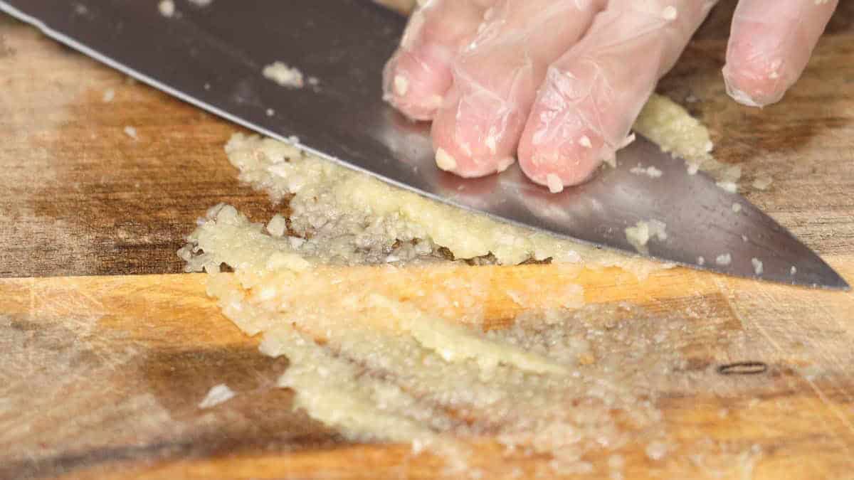Mashing the garlic into a paste-like consistency using the flat side of the knife.