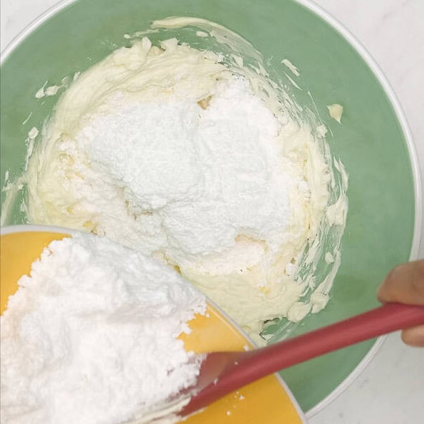 Mixing the cream cheese mixture using a hand mixer. Adding powdered sugar to the cream cheese mixture.