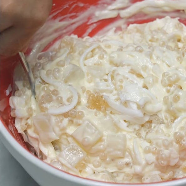 Adding the cream sauce to the bowl coconut strips, nata de coco, and sago. Mixing the salad mixture in the bowl.