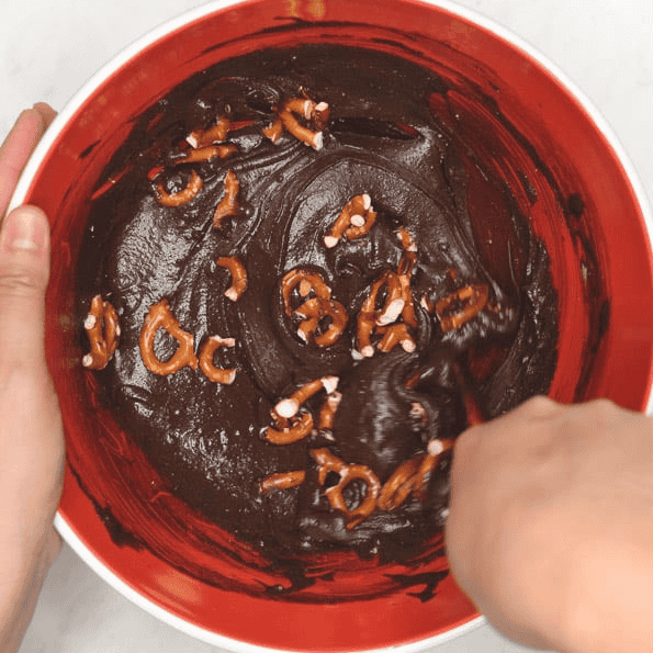 Mixing pretzel pieces into brownie batter in a red bowl.
