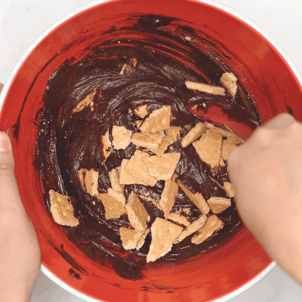 Mixing graham crackers into brownie batter in a red bowl.